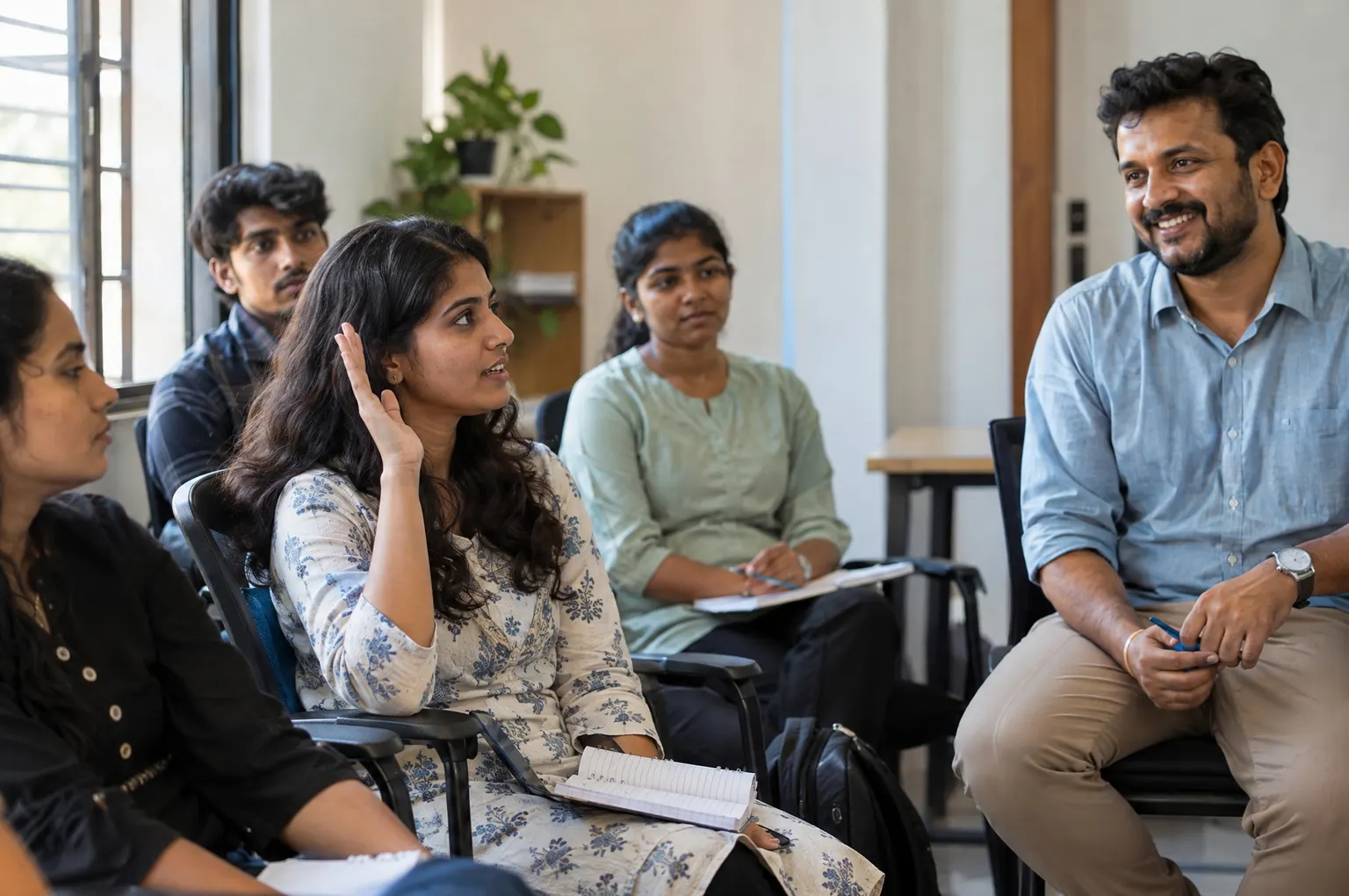 A young woman raising her hand to ask a question in a small workshop while a mentor smiles