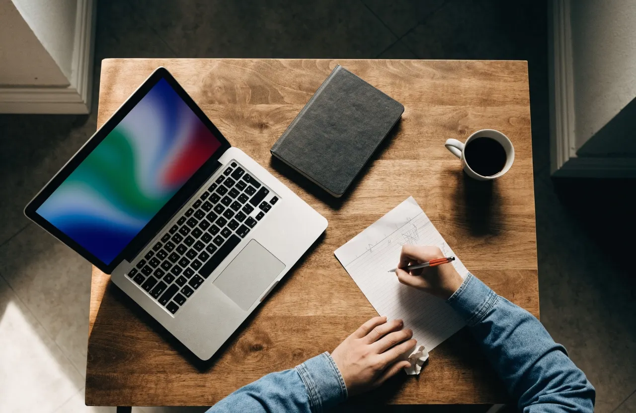 A laptop, a notebook and a hand taking notes on a wooden desk